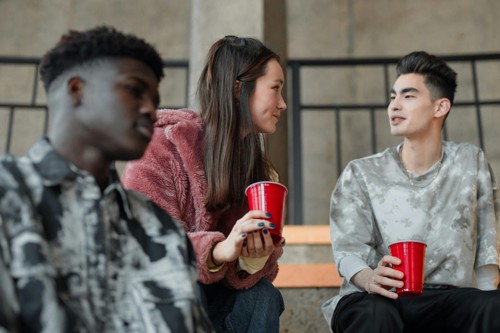 Three students, one girl and two boys, talk at university holding red cups.