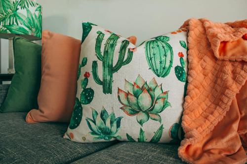 Un cojín de un verde intenso con motivos de cactus descansa sobre un sofá verde, junto a una manta y un cojín naranjas.
