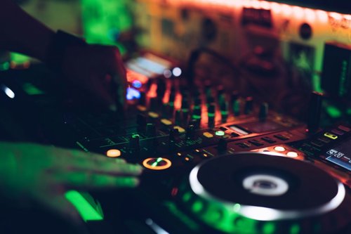Two hands work a DJ set under green and orange lights at a club.