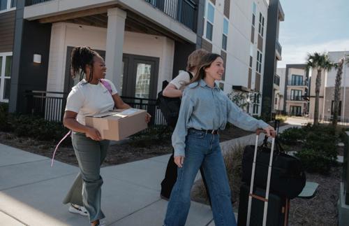 Three smiling students walk down a sunny suburban street with suitcases and boxes, excited to move in to their university accommodation.
