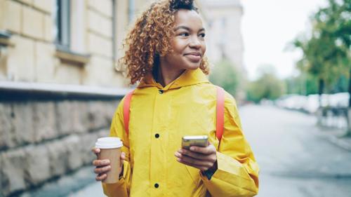 Una ragazza sorridente con un cappotto giallo cammina lungo la strada, con un caffè in una mano e il cellulare nell'altra.