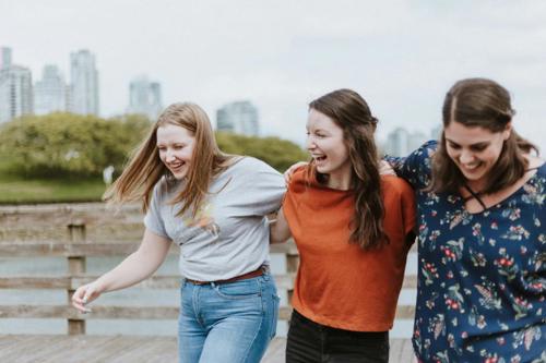 Three women laugh and walk arm-in-arm outside with a city skyline in the distance.