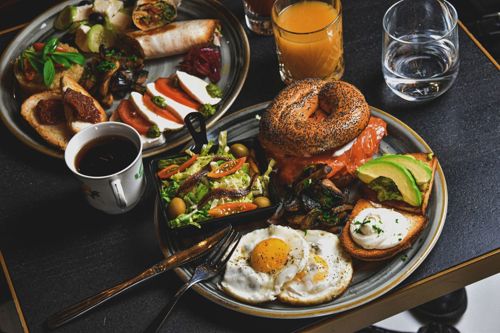 Two plates of bagels, eggs, and avocado sit beside cups of coffee and orange juice.